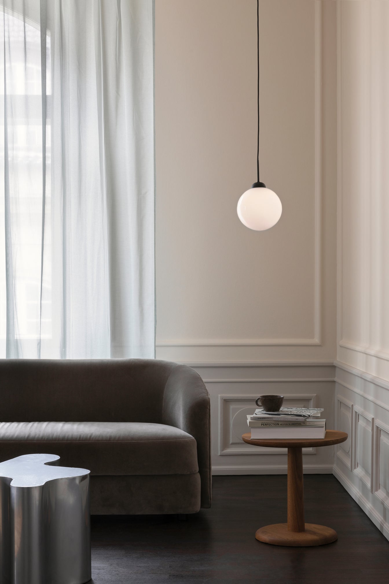 Pendel bestående af en rund skærm af opalglas og sort fatning samt ophæng, hængende ved sofa i dagligstuemiljø