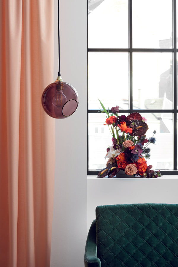 Rund lampe i lilla glas med et afskåret stykke med guldkant og kig til pæren. Fatning og rosetten er guldfarvet og ledningen er af sort stof.