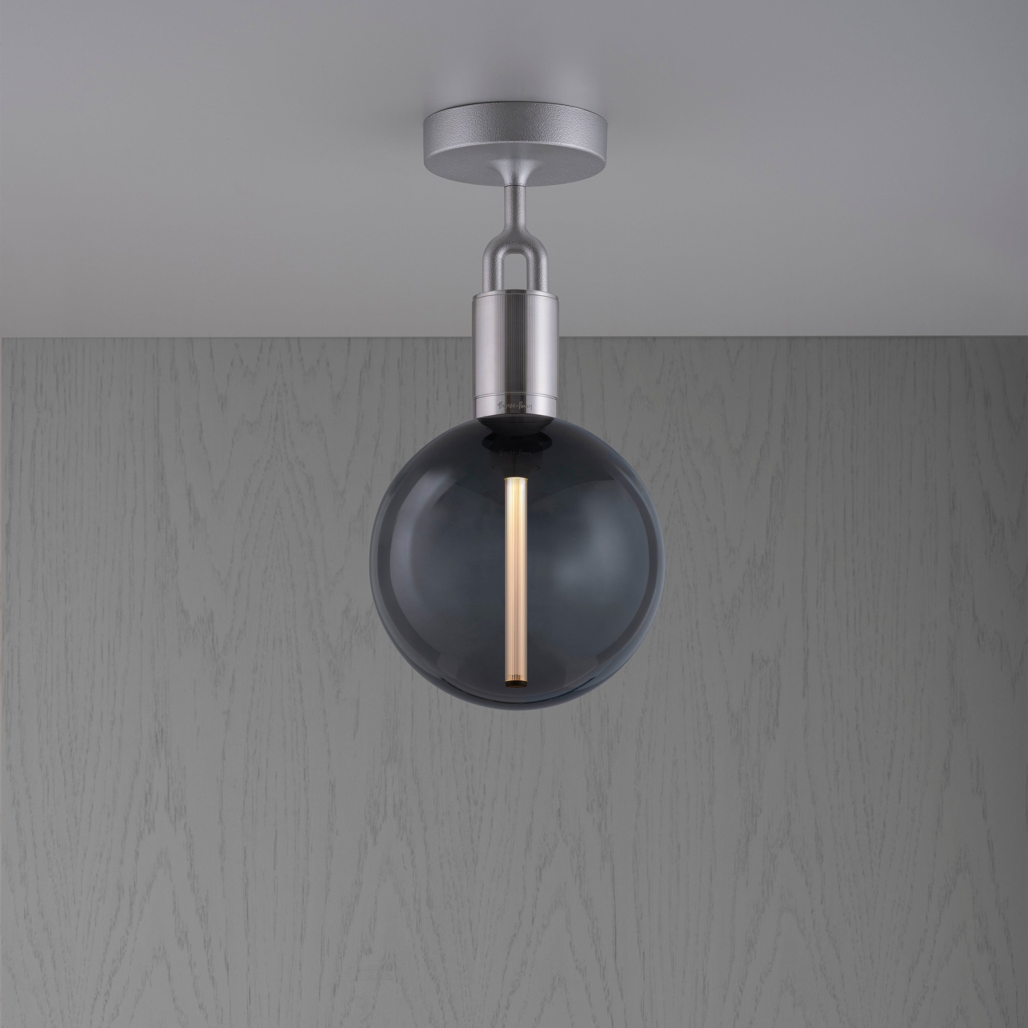 Loftlampe med rund skærm af røgfarvet glas og fatning i stål, på baggrund af gråt træ