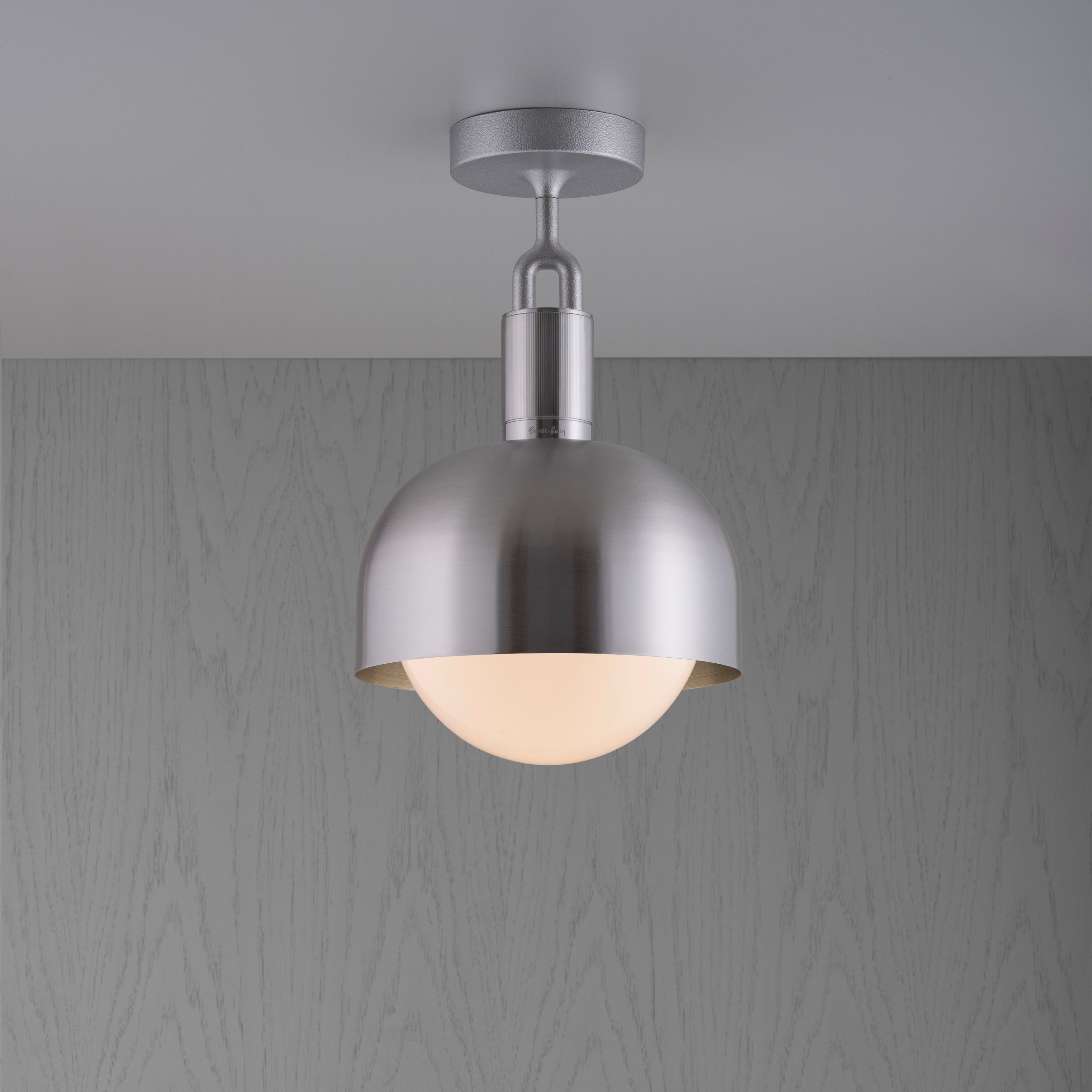 Loftlampe med fatning og skærm af stål samt pære i opalglas, på grå baggrund.