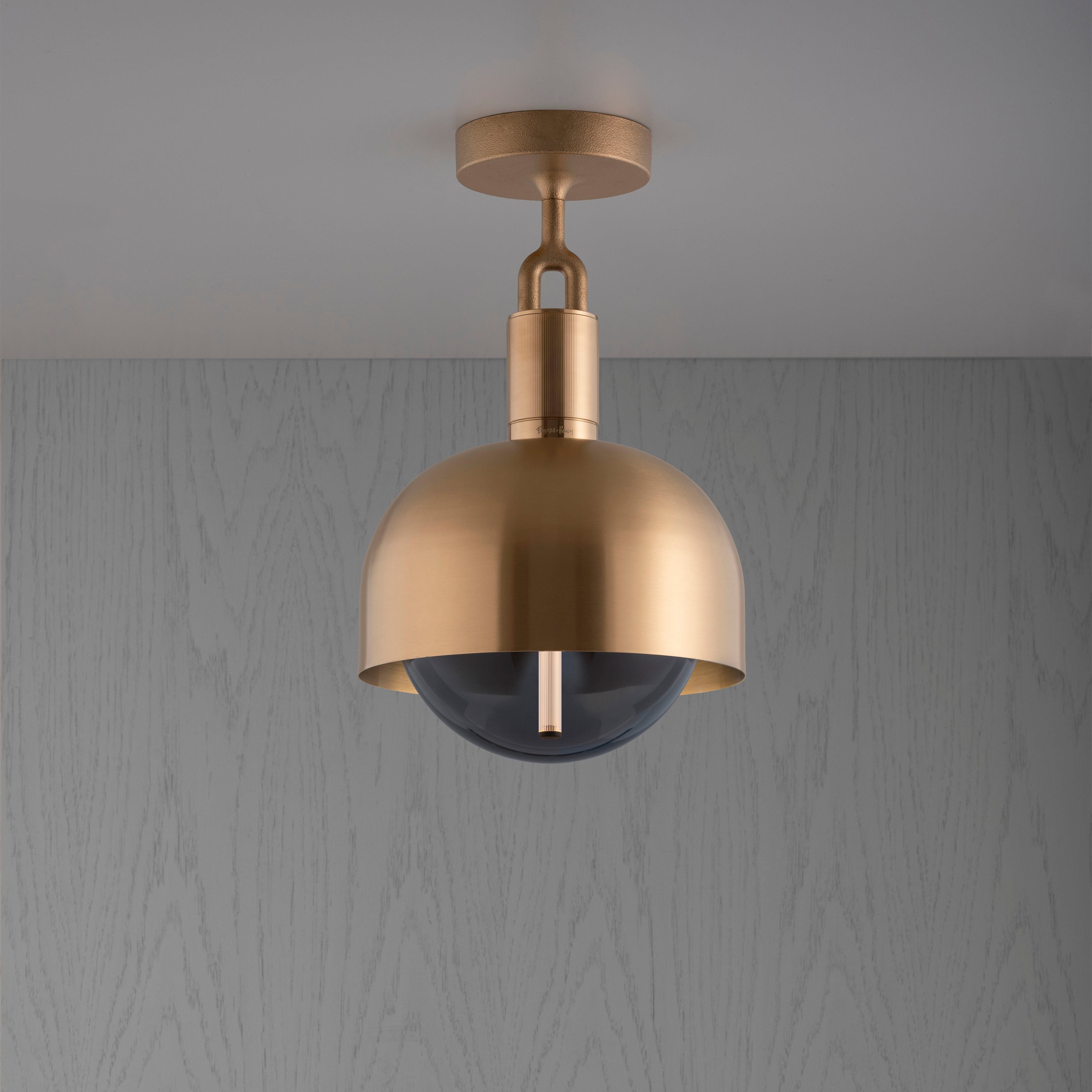 Loftlampe med fatning og skærm af messing samt pære af røgfarvet glas, på grå baggrund.