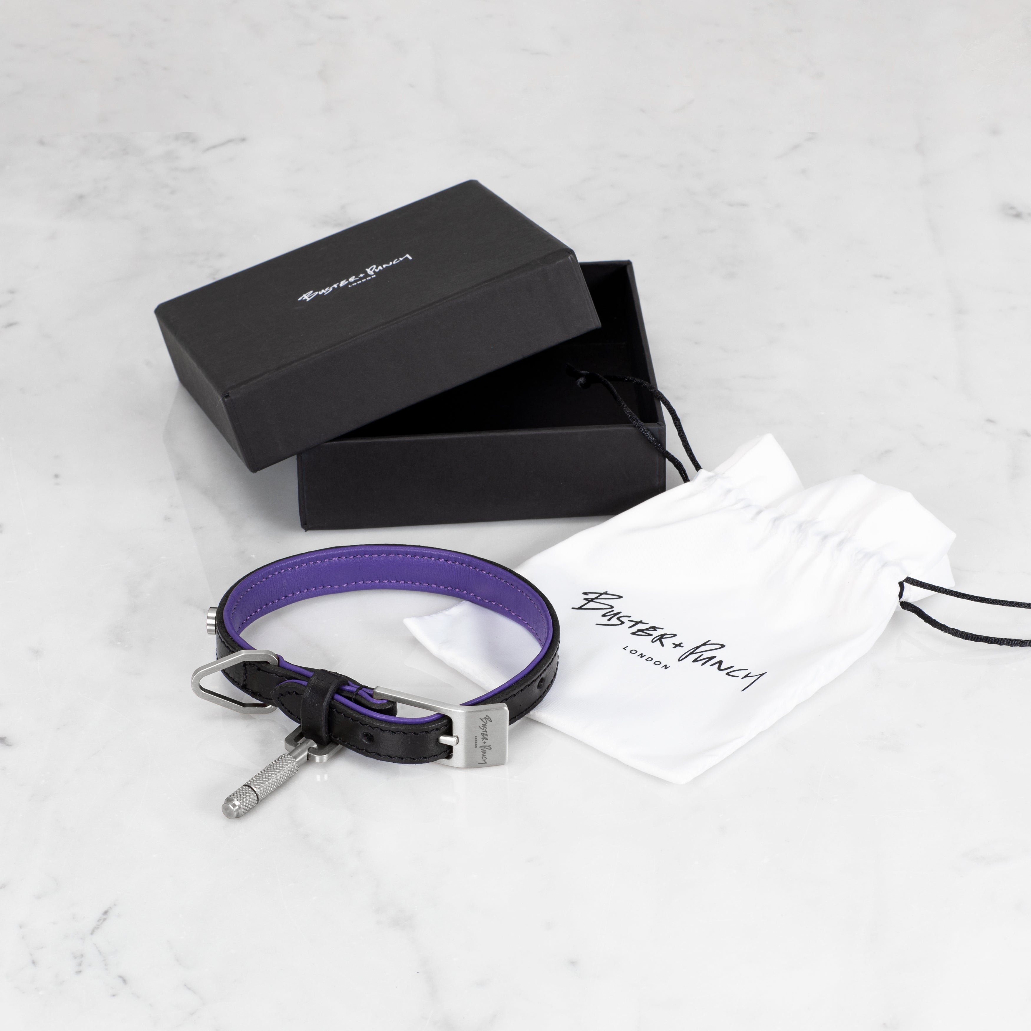 Æske indeholdende hundehalsbånd i sort og lilla læder med detaljer i stål, på hvid baggrund.