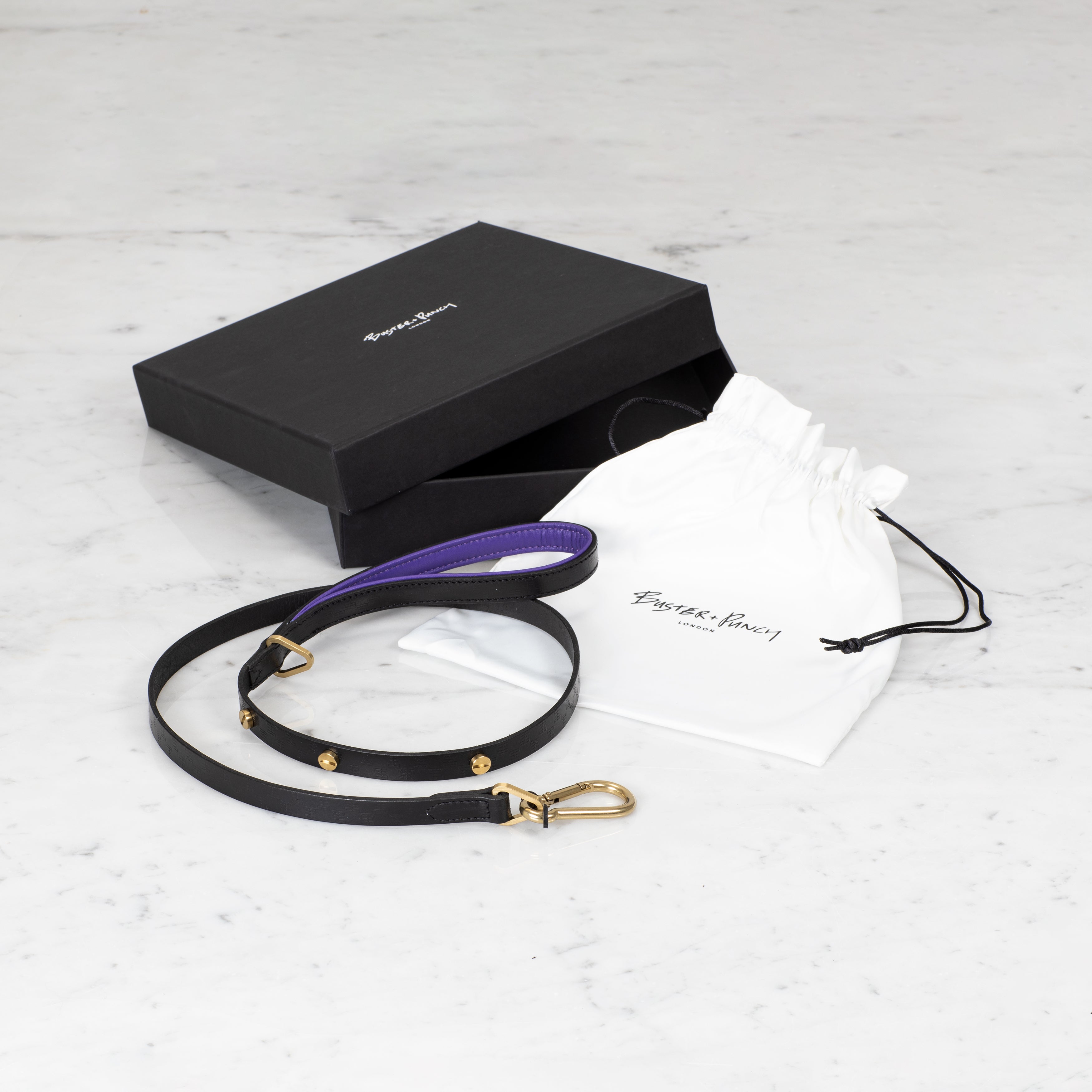 Æske indeholdende hundesnor i sort og lilla læder med detaljer i messing, på hvid baggrund.