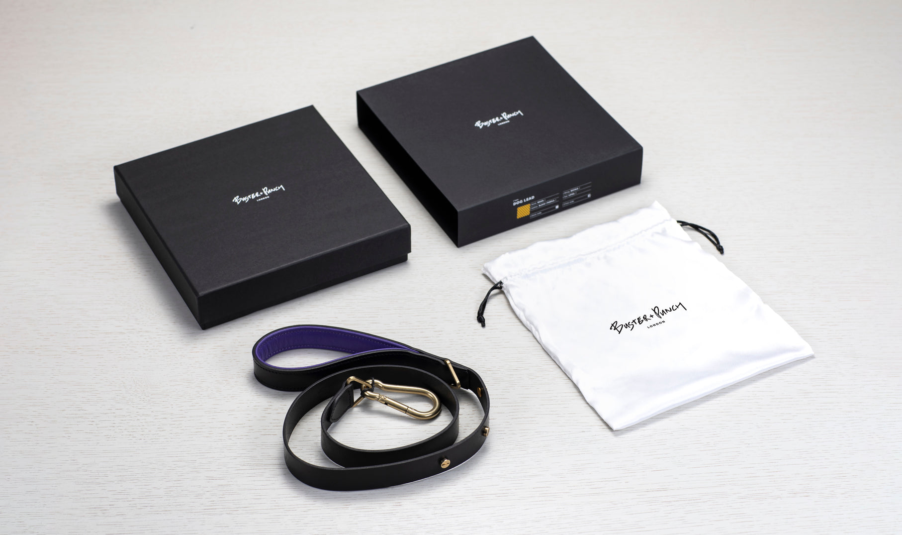Æske indeholdende hundesnor i sort og lilla læder med detaljer i messing, på hvid baggrund.