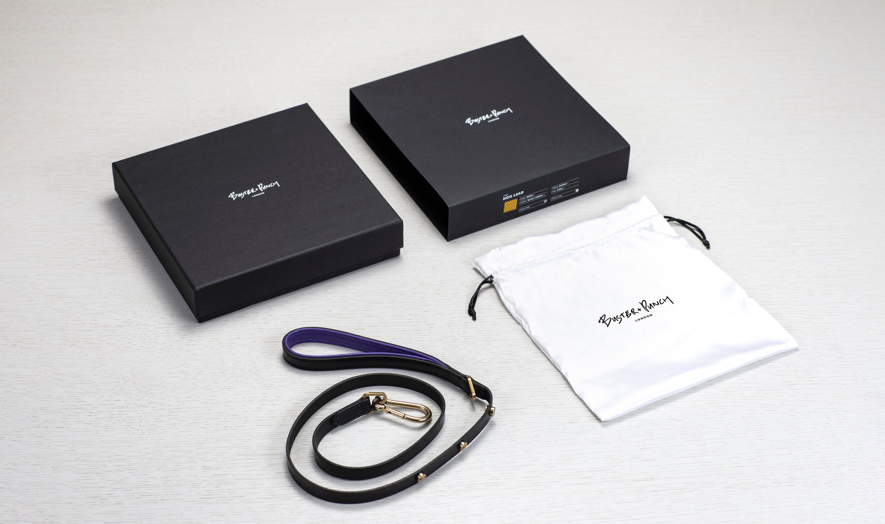 Æske indeholdende hundesnor i sort og lilla læder med detaljer i messing, på hvid baggrund.