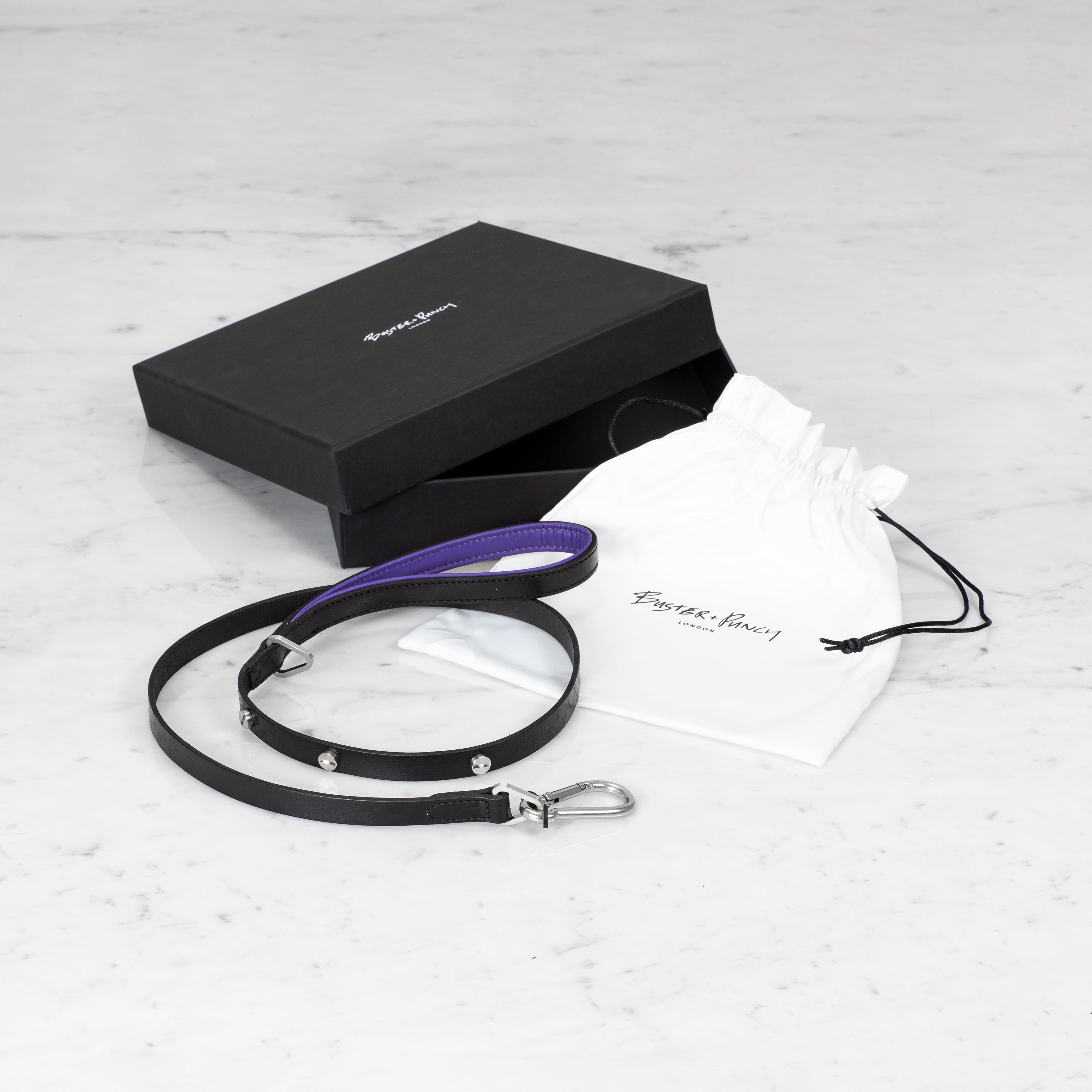 Æske indeholdende hundesnor i sort og lilla læder med detaljer i stål, på hvid baggrund.