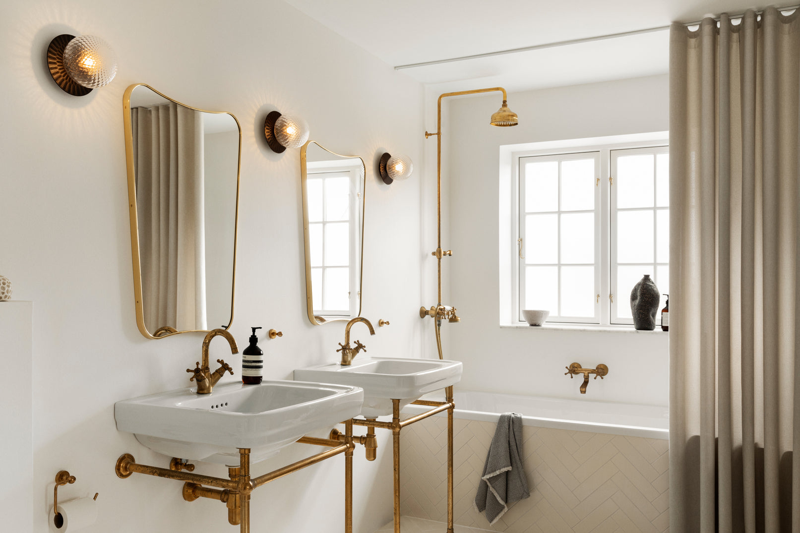 Tre væglampe med lampeskærme af klart optikglas og gylden fatning, hængende ved spejl på badeværelse