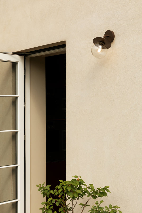 Væglampe med rund skærm af klart optikglas og bronzefarvet stel, på lys mur ved siden af dør