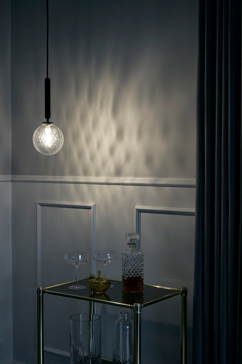 Pendel med rund skærm af klart optikglas og aflang grå fatning, i dagligstuemiljø