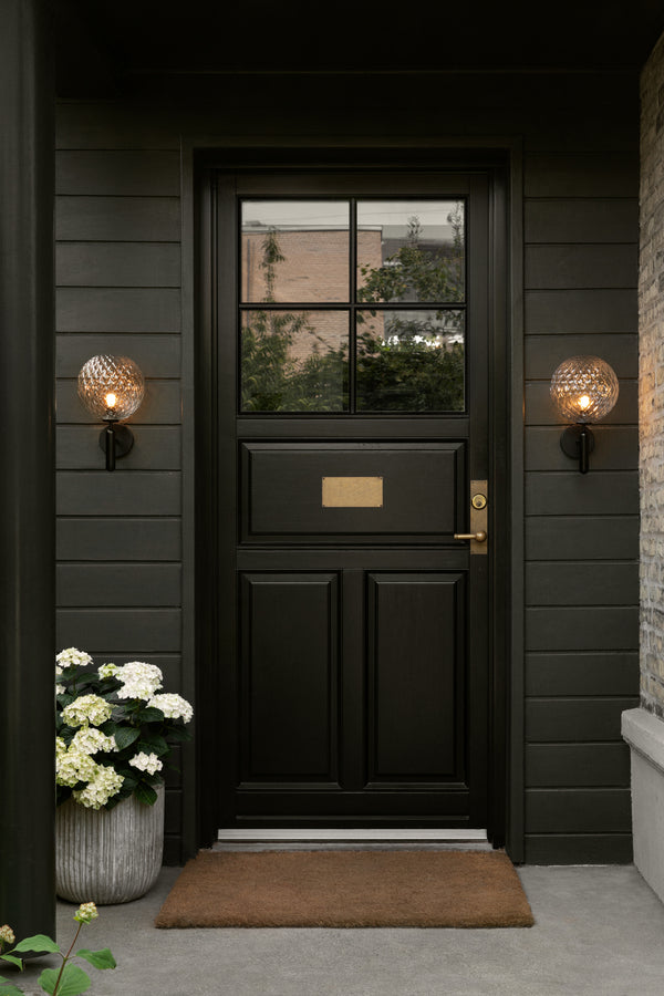 To væglampe med rund skærm af klart optikglas og sort stel, ved siden af hoveddør