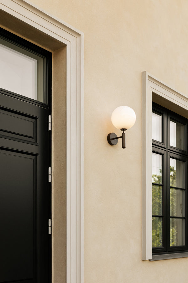 Væglampe med rund skærm af opalglas og bruneret stel, på husmur ved siden af hoveddør