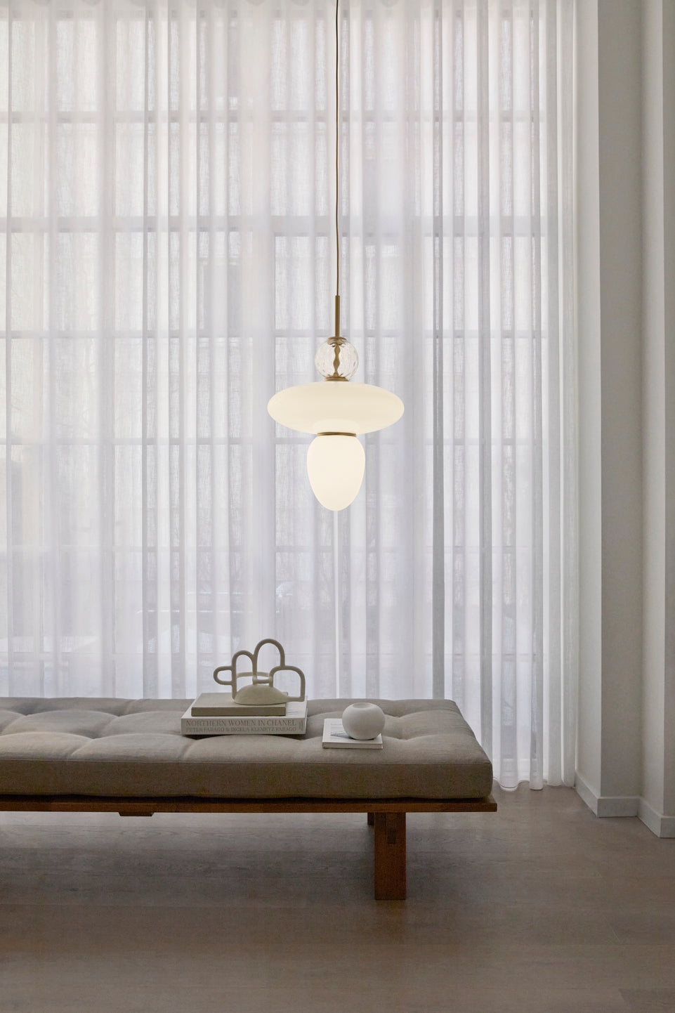Pendel bestående af tre glasskærme, én i klart optikglas og to i opalglas samt gyldent ophæng, hængende over daybed i dagligstuemiljø