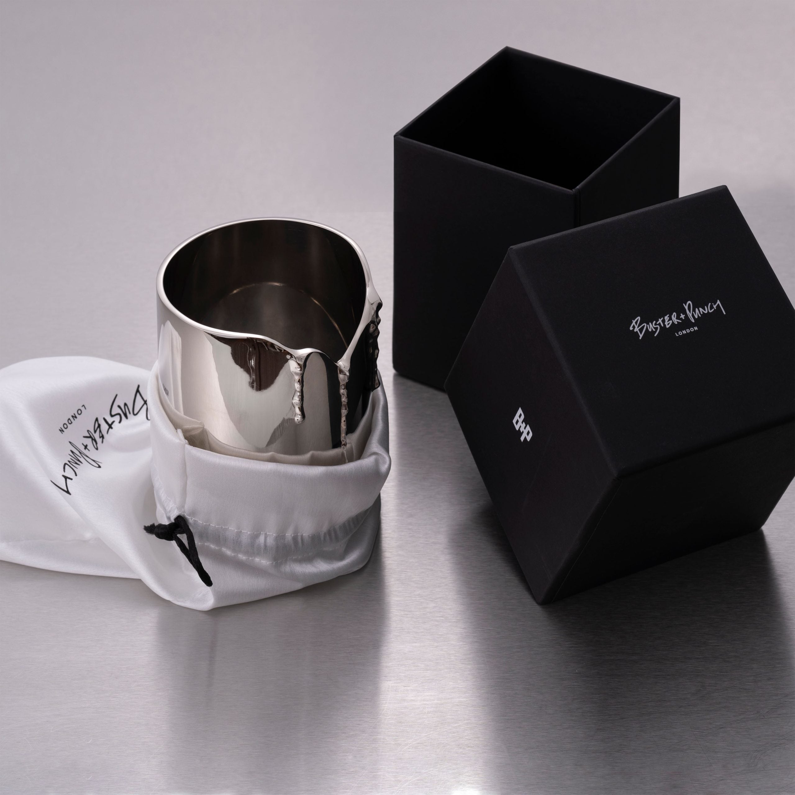 Æske indeholdende lystage i stål med duftlys i glas, på grå baggrund.