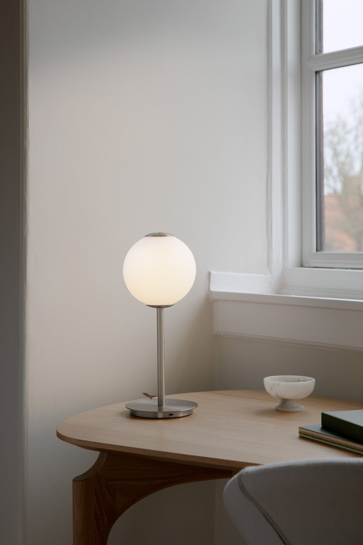 Audrey Table Tischleuchte aus Stahl mit leuchtendem Kugelschirm auf einem Holztisch am Fenster.