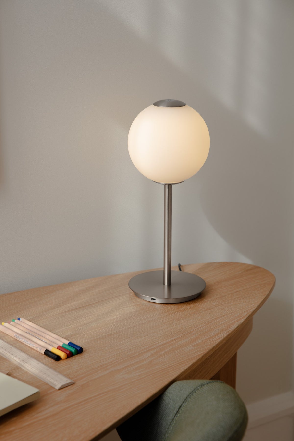Eingeschaltete Audrey Table Tischleuchte aus Stahl von UMAGE auf einem Holztisch mit Buntstiften.