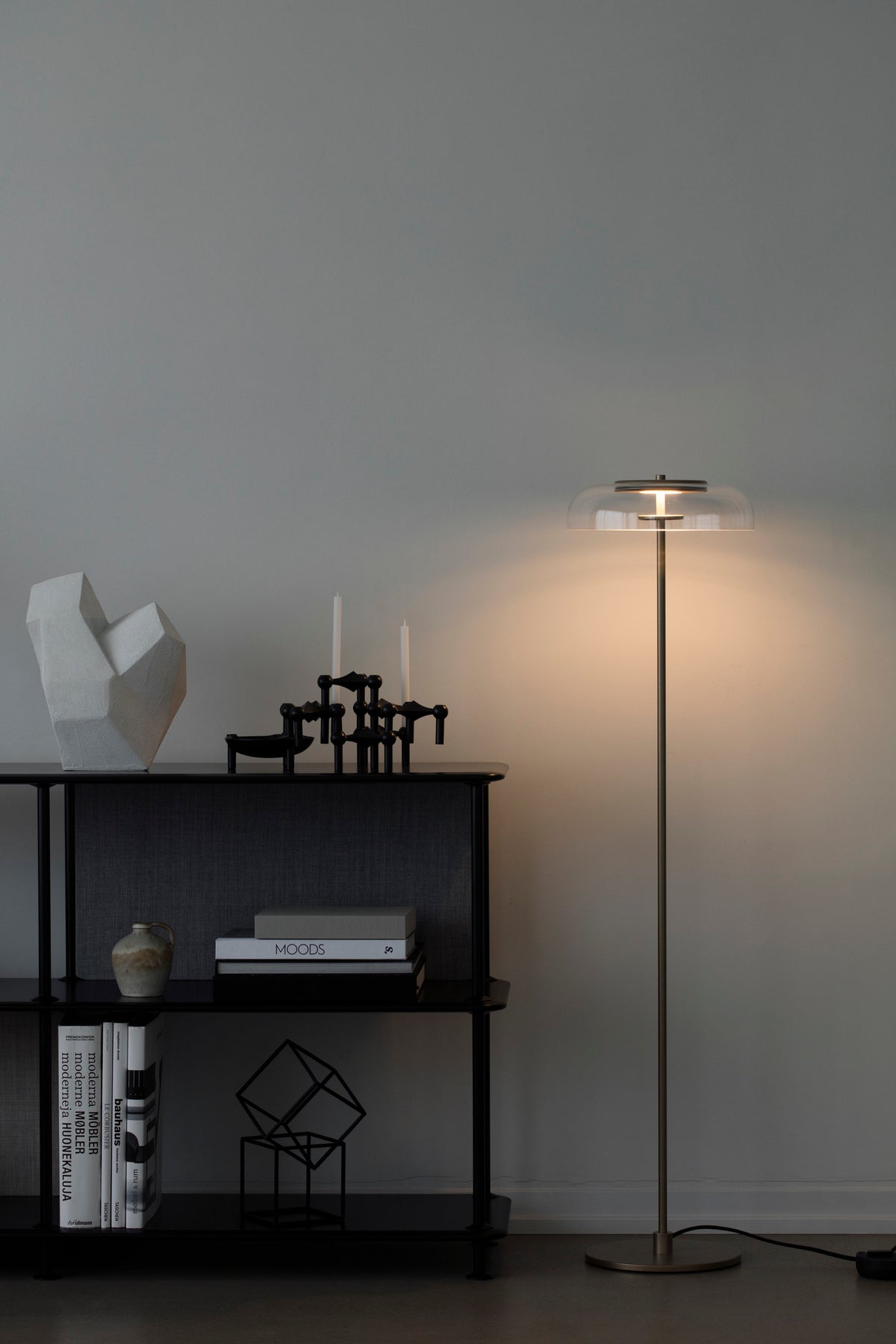 Gulvlampe med gyldent stel og fatning samt klar glasskærm, i dagligstuemiljø ved siden af reol