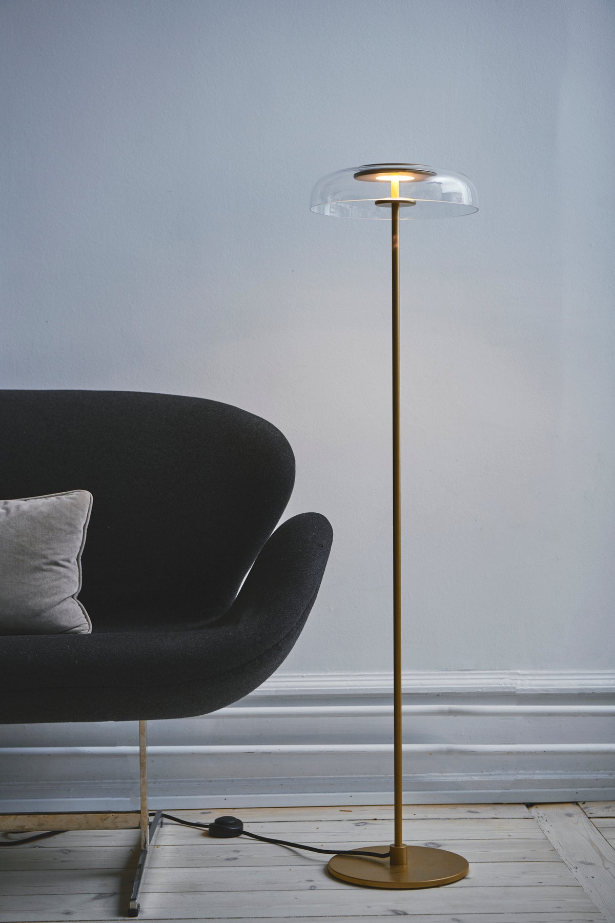 Gulvlampe med gyldent stel og fatning samt klar glasskærm, i dagligstuemiljø
