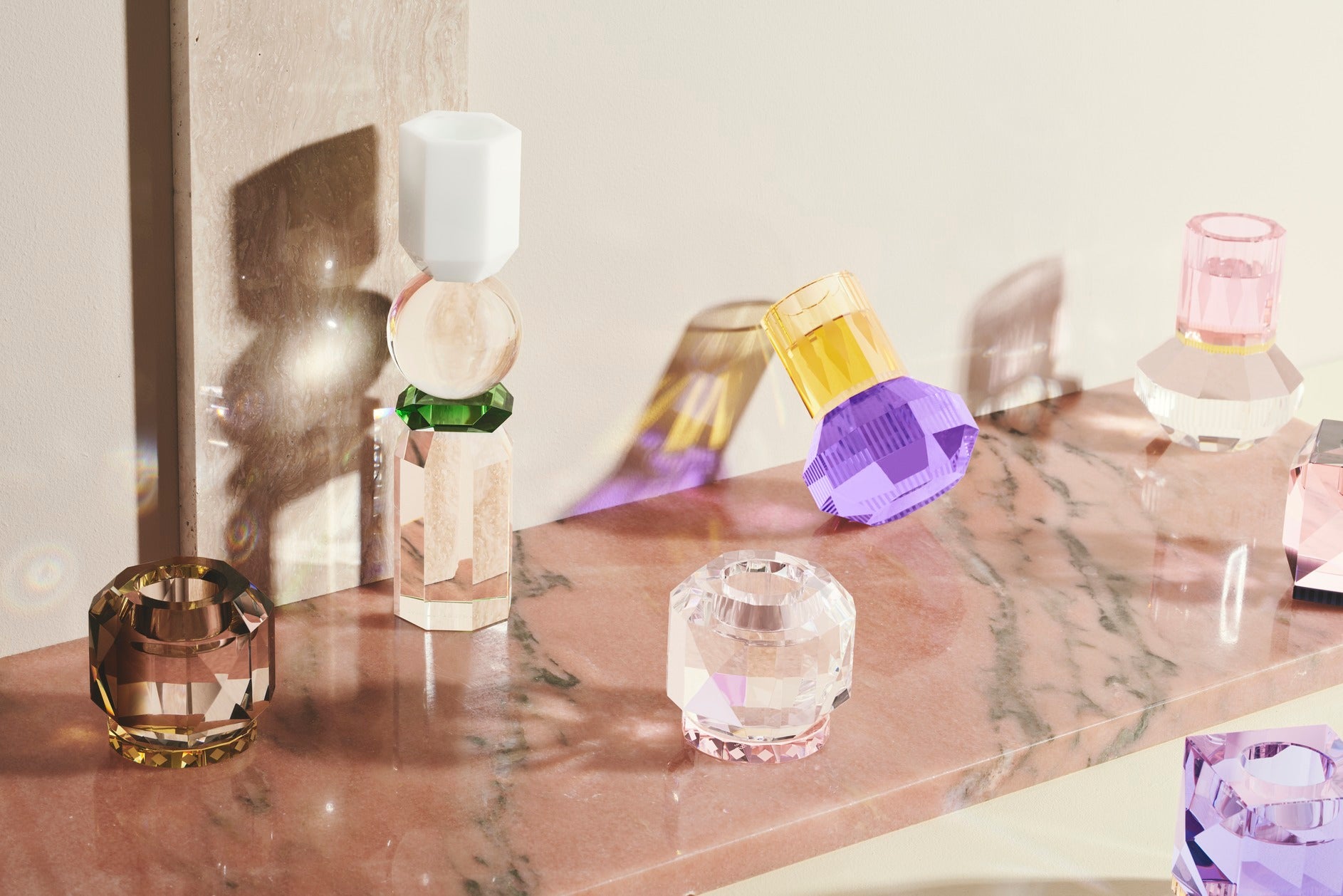 Opstilling af forskellige fyrfadslysestager i farvet krystalglas, placeret på laksefarvet marmorplade.
