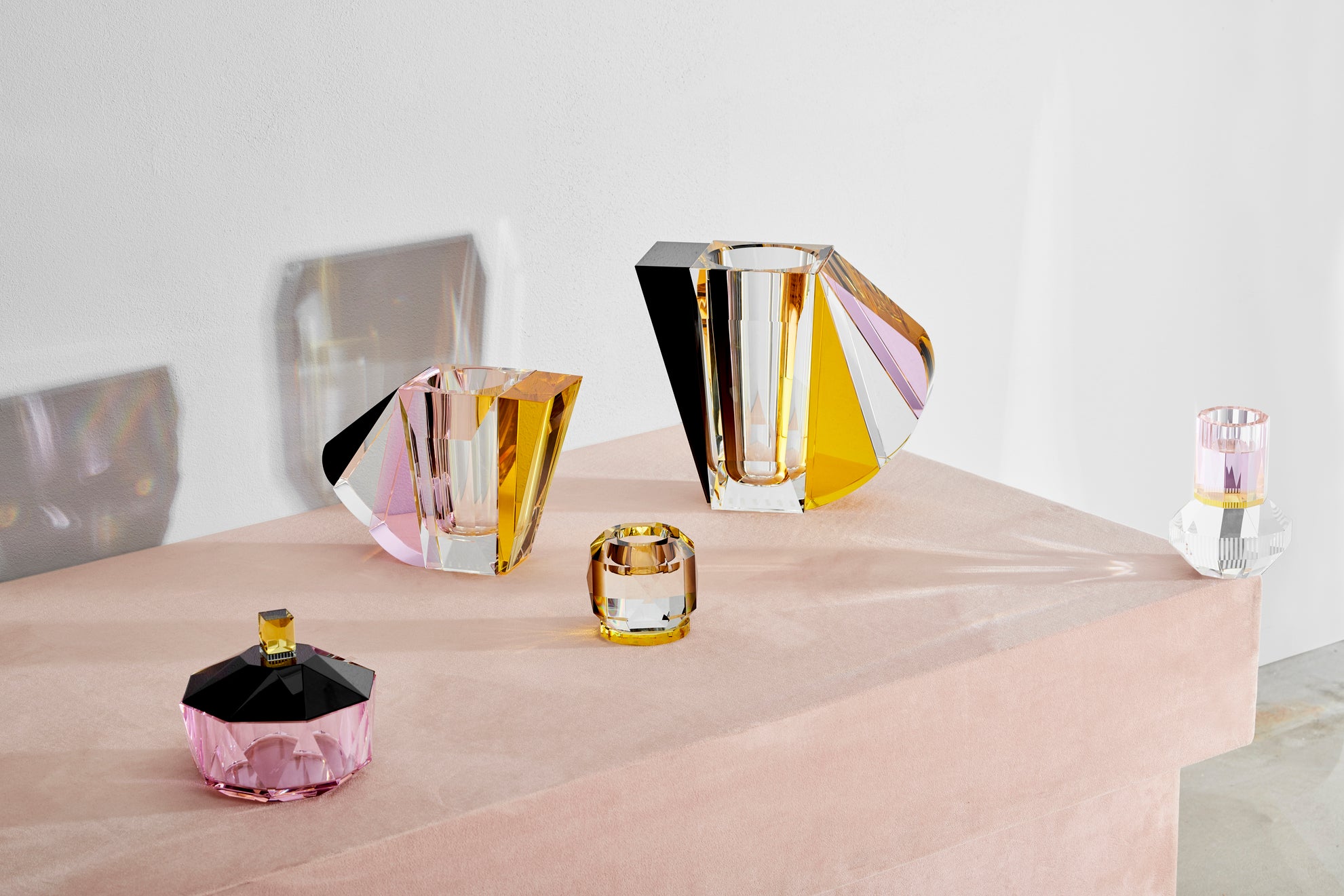 Opsætning af vaser, lysestager samt bonbonniere i farvet krystal, på lyserødt bord og med hvid baggrund.