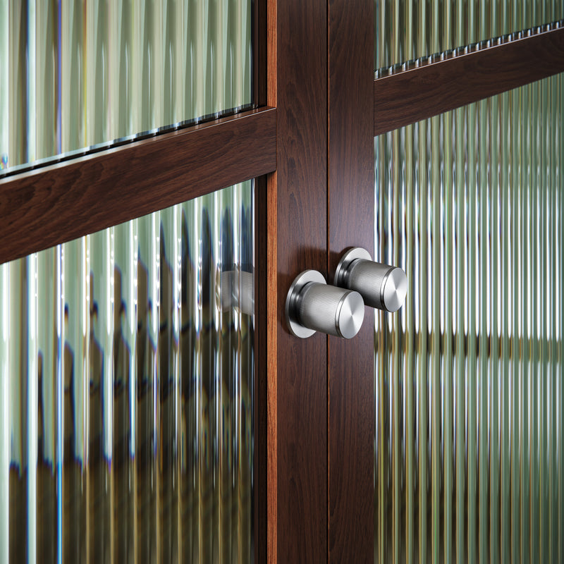 Eksklusiv dørknop i rustfri stål med linjeret mønster. Her ses to dørknopper monteret på en trædør med glasrude.