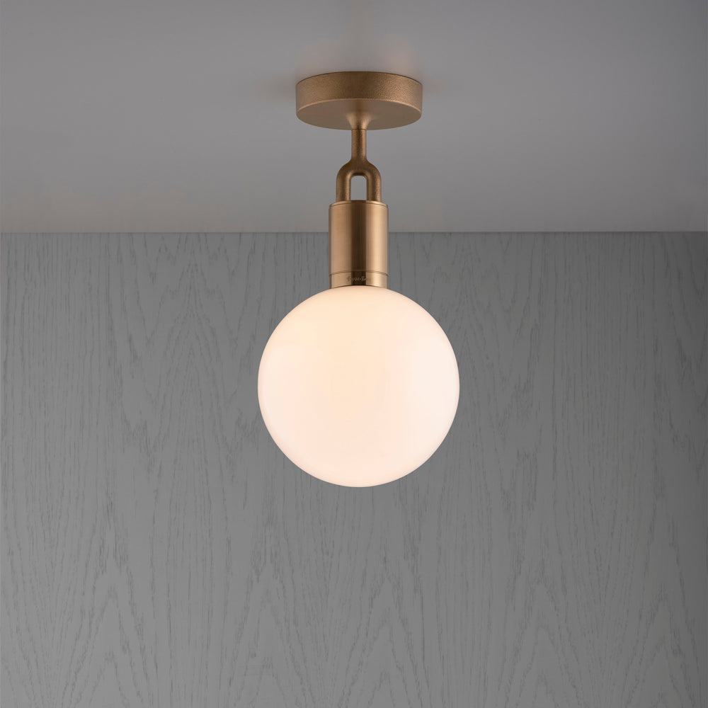 Loftlampe med rund skærm af opalglas og med fatning i messing, på baggrund af gråt træ.