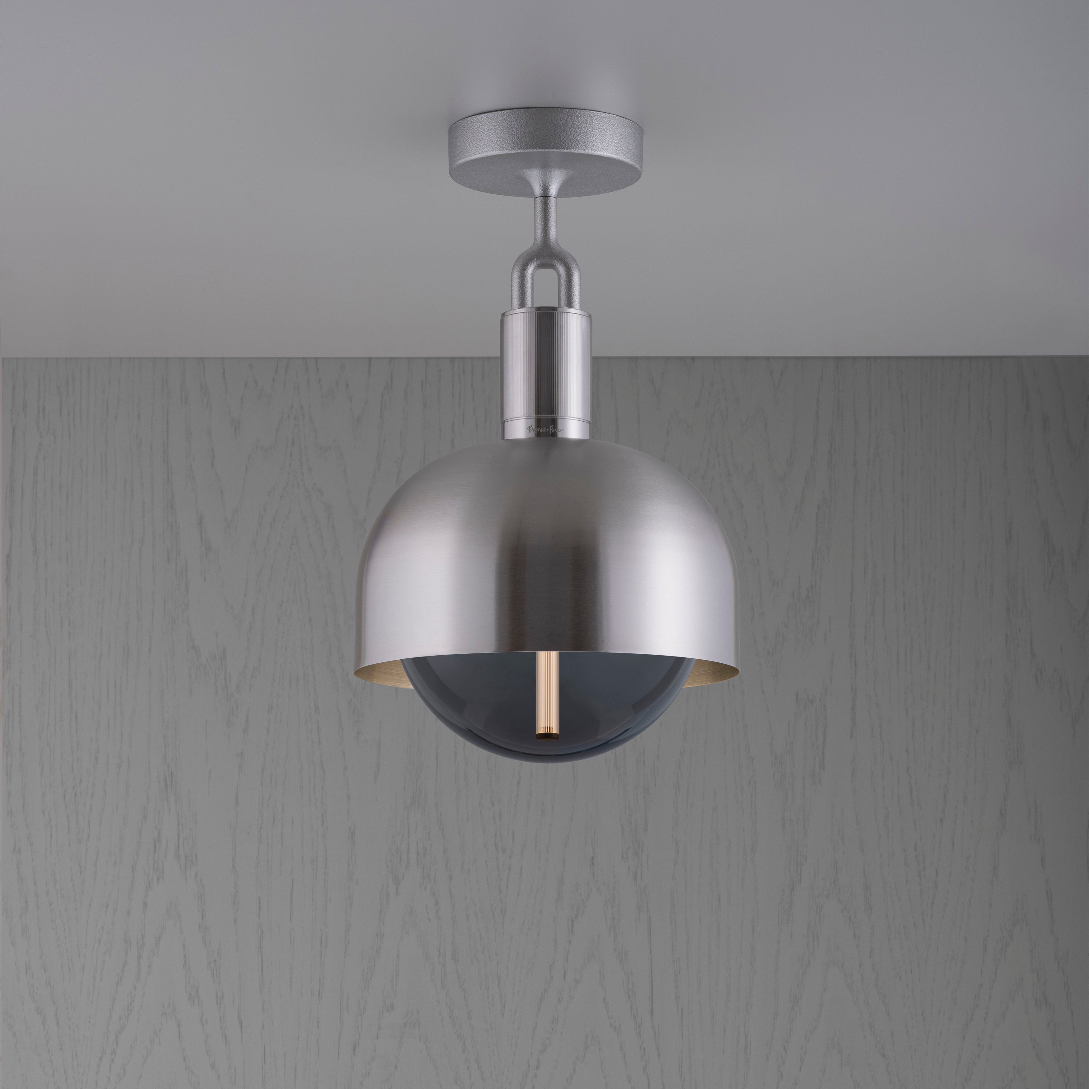 Loftlampe med fatning og skærm af stål samt pære af røgfarvet glas, på grå baggrund.