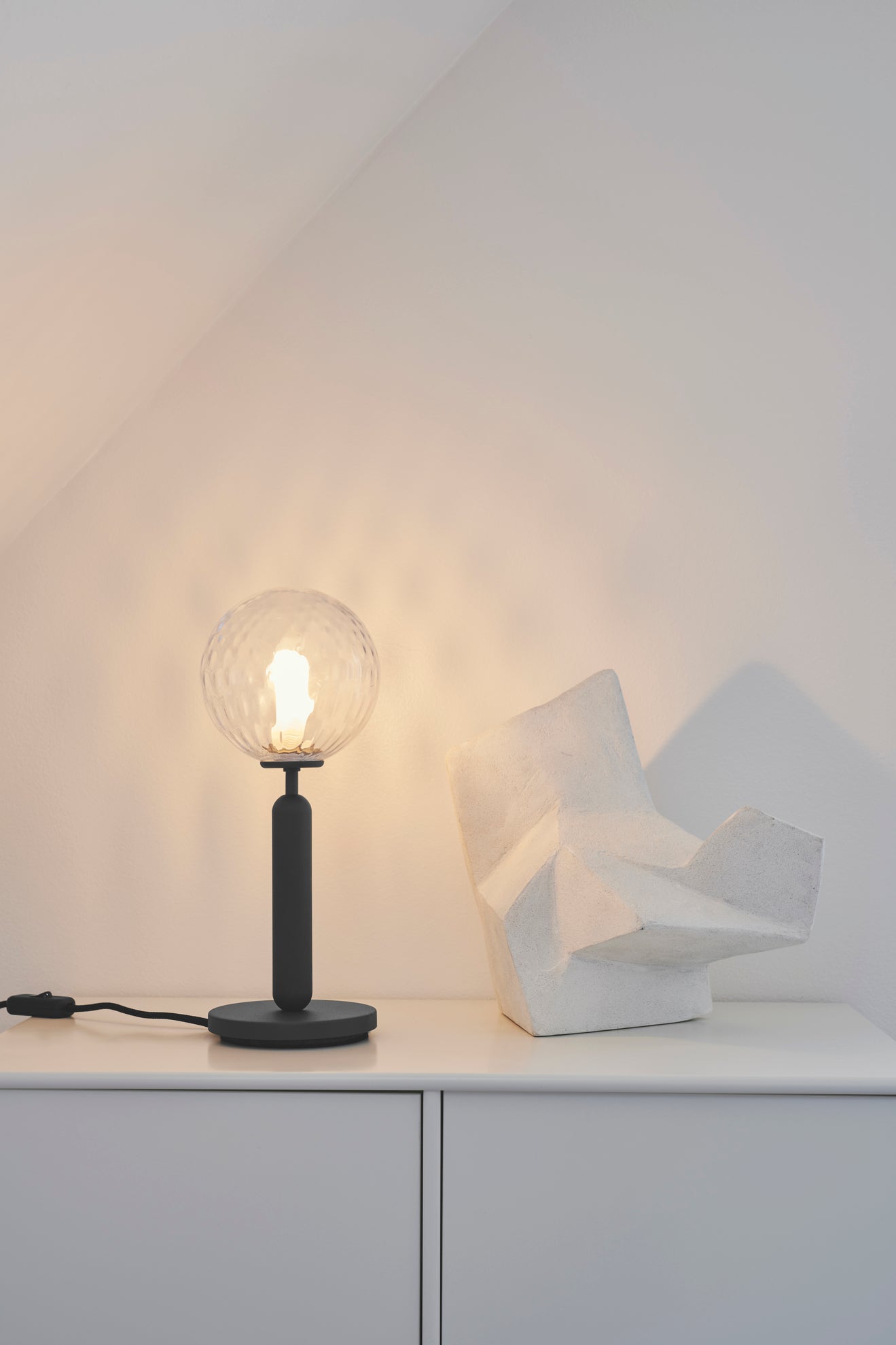 Bordlampe med rund skærm af klart optikglas og mørkegråt stel, i dagligstuemiljø