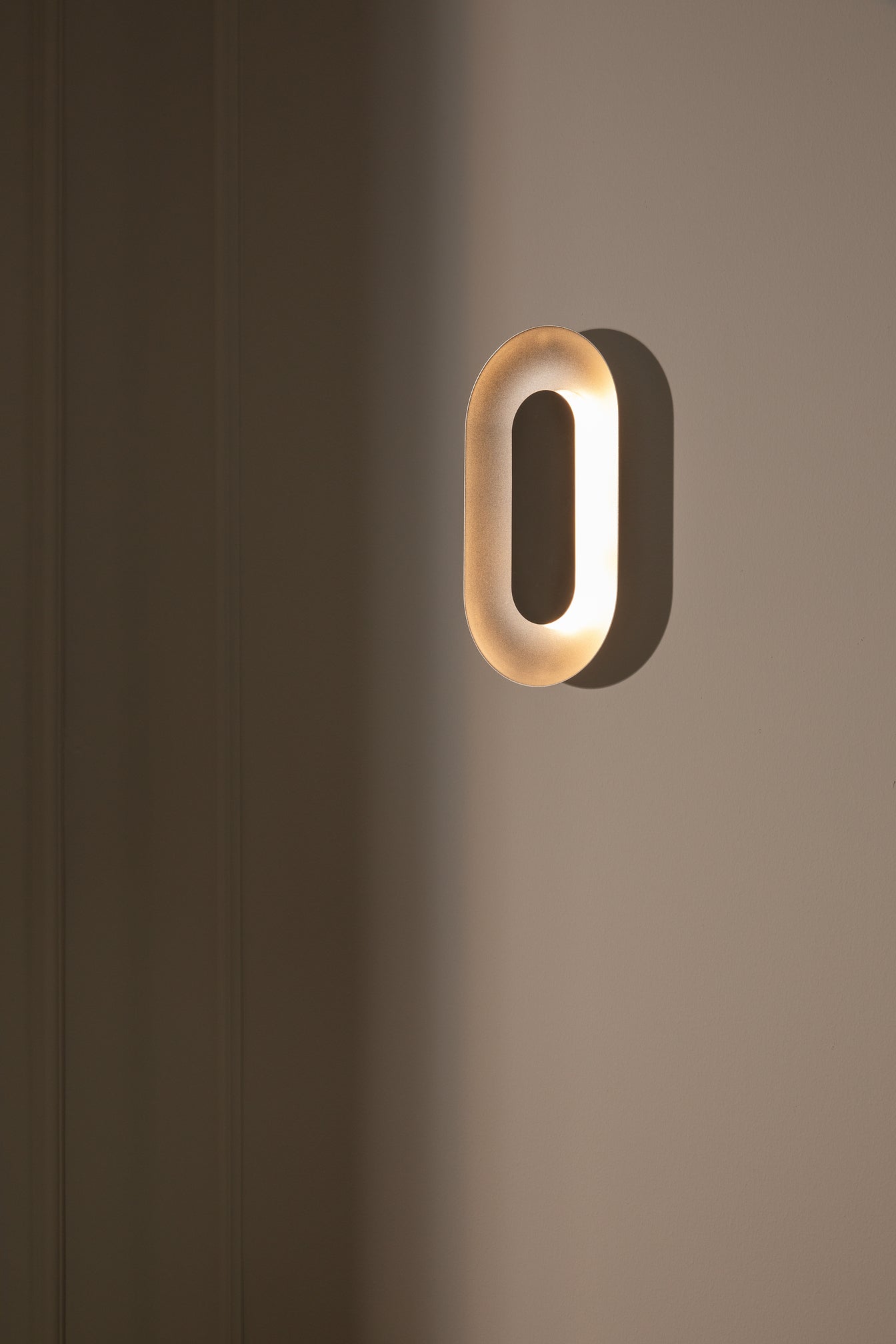 Oval sølvfarvet væglampe med indirekte lyskilde, på hvid væg