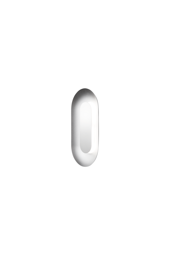 Oval sølvfarvet væglampe med indirekte lyskilde, på hvid baggrund