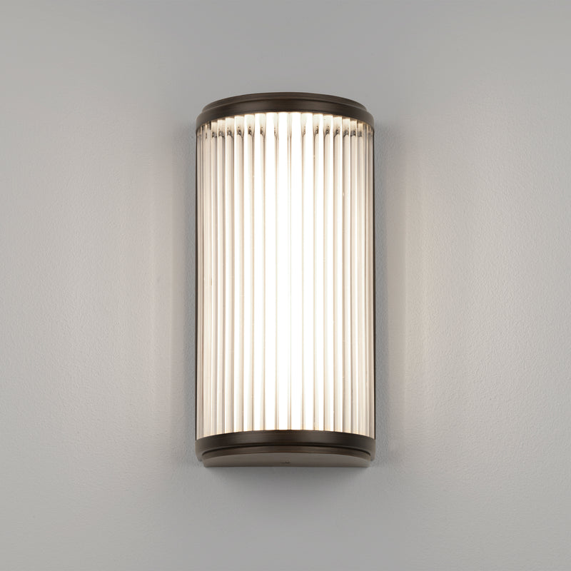 Lampe med top, bund og bagside i bronzefarvet stål samt en glasskærm med nedadgående linjer.